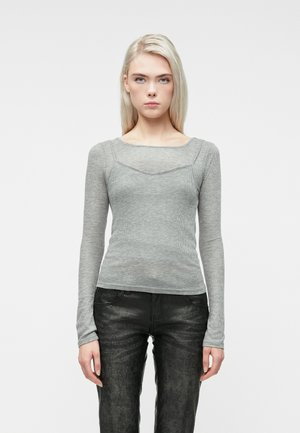 Młoda kobieta z długimi blond włosami, ubrana w dopasowaną szaro-skalistą bluzkę z długim rękawem i czarne teksturowane spodnie, stojąca na tle białego tła.