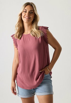 Hymyilevä nainen, jolla on vaaleat hiukset, pukeutunut hiuksetön mauve-väriseen, pitsikoristeltuun toppiin ja vaaleisiin farkkushortseihin, seisoo yksinkertaisen taustan edessä.