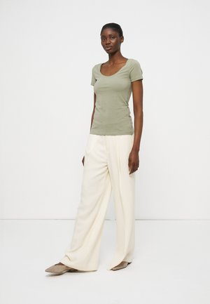 Žena stojaca, oblečená v svetlozelenom tričku s krátkym rukávom a výstrihom do U, širokých bielych nohaviciach a béžových špicatých topánkach na bielom pozadí.