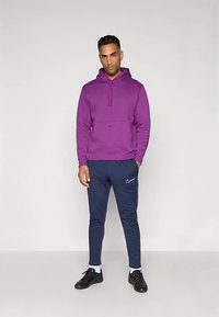 Muž stojící v fialové mikině, tmavě modrých Nike teplácích, bílých ponožkách a černých teniskách na jednoduchém světle šedém pozadí.