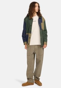 Ein mehrfarbiger Patchwork-Jacke in Grün, Marineblau und Beige, mit einem Kragen und Knopfverschluss, kombiniert mit beigen, lässigen Hosen und braunen Schuhen.