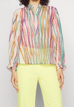 Žena v průsvitné, vícebarevně pruhované blůze s řasenými manžetami a světle žlutých kalhotách, stojící před jednobarevným pozadím.