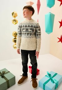 Knit sweater with white, grey, and black patterns, dark jeans, and brown suede sneakers. Surrounded by colorful geometric decorations and gift boxes.