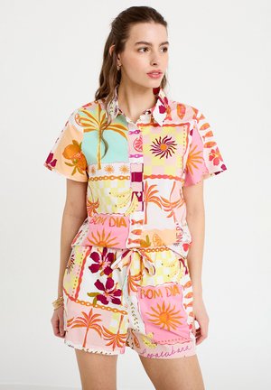 Kvinde iført farverig, ensartet kortærmet skjorte med knapper og shorts med tropiske motiver og ord som "Bom Dia."