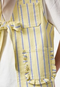 Gilet sans manches rayé jaune et bleu avec un lien à l’avant, comprenant une poche simple et une poche à volants, porté sur une chemise blanche.