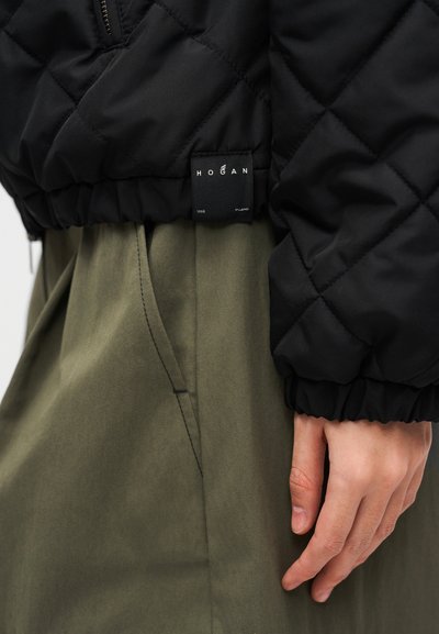 Μαύρο καπιτονέ μπουφάν με ριμπ μανσέτες, εμφανής ετικέτα. Φοριέται πάνω από χακί παντελόνια με πλαϊνές τσέπες, προβάλλοντας κομψό σχεδιασμό και υλικά.