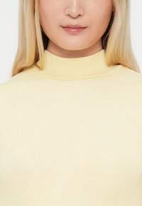 Keltainen mock turtleneck -paita, joka on valmistettu joustavasta kankaasta. Siinä on sileä pinta ja tiivis istuvuus. Minimalistinen tyyli ilman kuvioita.