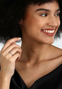 Silberner Ring mit kleinen klaren Steinen, am Zeigefinger getragen, neben einem Model mit dunklem, lockigem Haar und einem schwarzen Satin-Oberteil.