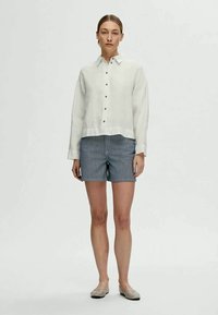 Kvinde står iført en hvid skjorte med knapper og lange ærmer, blå og hvide stribede shorts samt beige flade sko foran en ensfarvet baggrund.