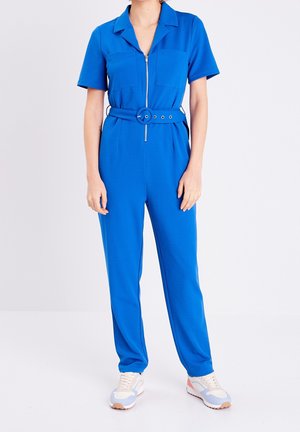 Combinaison courte bleue à manches courtes avec fermeture éclair à l'avant, col, deux poches poitrine, une taille ceinturée et une coupe décontractée. Associée à des baskets de couleur claire.