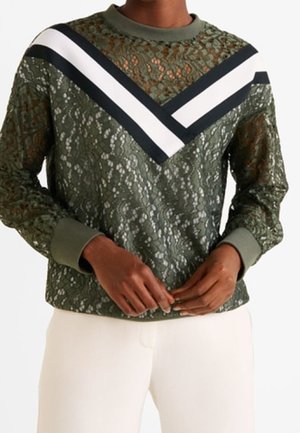 Donkergroene kanten top met lange mouwen en zwart-witte chevronstrepen op de borst, gedragen met gebroken-witte broek, handen losjes in elkaar gevouwen voor het lichaam.