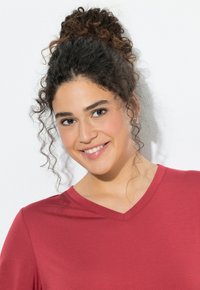 Bordowa koszulka z dekoltem w serek, z krótkimi rękawami i miękką, gładką fakturą. Modelka ma kręcone włosy upięte w kok i delikatny uśmiech.