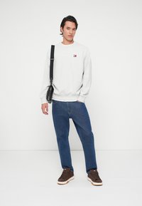 Άνδρας φορεμένος με ανοιχτό γκρι φούτερ, μπλε τζιν, καφέ αθλητικά παπούτσια και μαύρη τσάντα ώμου, στέκεται μπροστά σε λευκό φόντο.