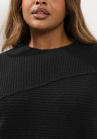 Svart ribbad stickad tröja med rund halsringning och diagonalt strukturerat mönster över bröstet, som framhäver det mjuka, tjocka tyget.