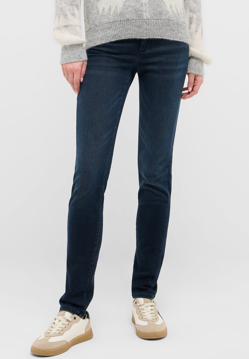 Tmavomodré úzke džínsy vyrobené z denimu, s hladkou textúrou a stredným pásom, doplnené béžovými a bielymi teniskami.