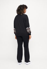 Svart sweatshirt med färgglada broderade mönster på ärmarna, parat med svarta byxor och vita sneakers. Modell sedd bakifrån.