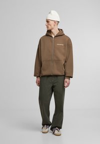 Brun zip-up hoodie med frontlommer og teksten "FORBUDT". Matchet med olivengrønne bukser og cremede sneakers. Hvid strikket beanie på hovedet.