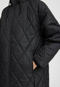 Manteau matelassé noir avec un motif en losanges, doté d'un col montant, d'une fermeture éclair et de poches latérales avec des zippes cachées.