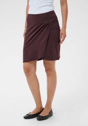 Femme portant une jupe portefeuille brun foncé à hauteur des genoux, un haut blanc et des ballerines noires, debout devant un fond uni.