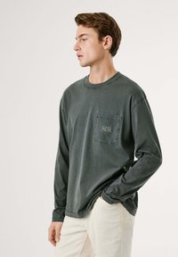 Camisa de manga larga de color verde oscuro, material de algodón, cuello redondo, bolsillo en el pecho con detalle del logo, combinada con pantalones de color claro.