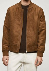 Veste bombardier en daim marron avec fermeture éclair à l'avant, poignets côtelés et finition lisse. Portée sur un pull foncé et un pantalon clair.