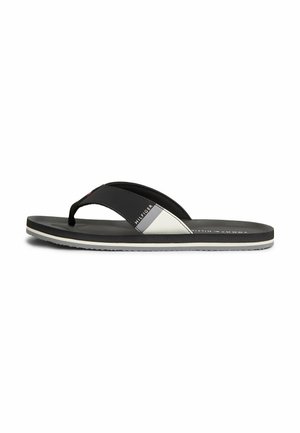 Schwarz-weiße Tommy Hilfiger Flip-Flop-Sandale mit einfachem Zehensteg und flacher Sohle, von der Seite auf weißem Hintergrund gezeigt.
