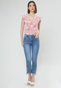 Blumenmuster pinke Bluse mit Rüschensleeves, kombiniert mit hellblauen Skinny Jeans mit Knopfakzenten an den Bündchen und weißen offenen Schuhen mit Absatz.