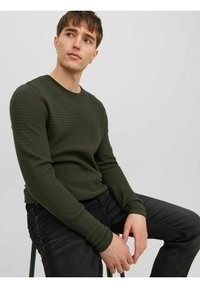 Oliven grøn, struktureret skjorte med lange ærmer, rund halsudskæring og tætsiddende manchetter, parret med mørke jeans. Sittende på en stol.
