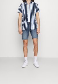 Camisa de botones de manga corta con rayas verticales en azul marino y blanco, combinada con pantalones cortos de mezclilla y zapatillas blancas, sobre un fondo neutro.