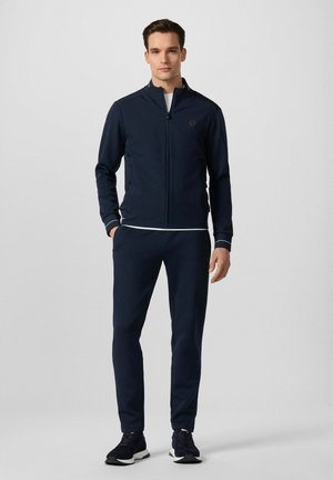 Hombre de pie con chaqueta azul marino con cremallera, pantalones a juego, camiseta interior blanca y zapatillas deportivas negras sobre un fondo claro y liso.