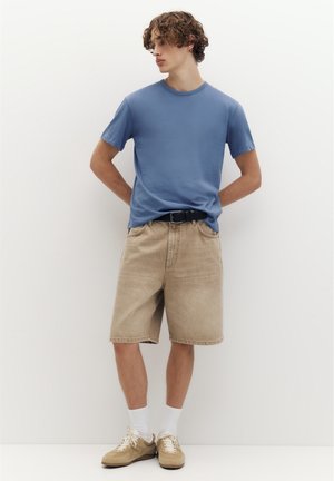 Jeune homme aux cheveux bouclés portant un t-shirt bleu, un short beige avec une ceinture noire, des chaussettes blanches et des baskets beiges, debout contre un mur blanc.