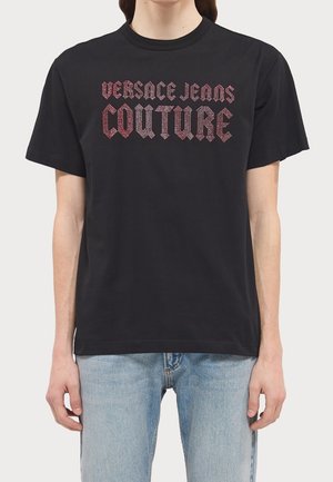 Personne portant un t-shirt noir à manches courtes avec l'inscription "Versace Jeans Couture" en lettres gothiques rouges, associé à un jean denim bleu clair.