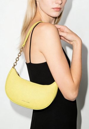 Blonde Frau in schwarzem Spaghettiträgerkleid, die eine gelbe, halbmondförmige Handtasche mit goldener Kette über der Schulter trägt.