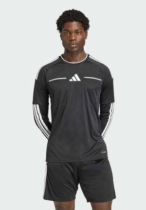 Junger Mann, der mit den Händen hinter dem Rücken steht, trägt ein schwarzes Adidas Langarm-Fußballtrikot und Shorts mit weißen Streifen und Logo.