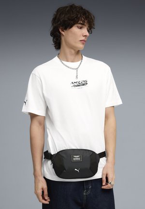 Banane noire et spacieuse avec un panneau en maille texturée, arborant un logo. Portée sur un t-shirt graphique blanc et associée à un jean foncé.