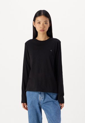 Camiseta negra de manga larga hecha de algodón suave, con cuello redondo y un pequeño logo negro en el lado izquierdo del pecho. Jeans de denim lavados a máquina.