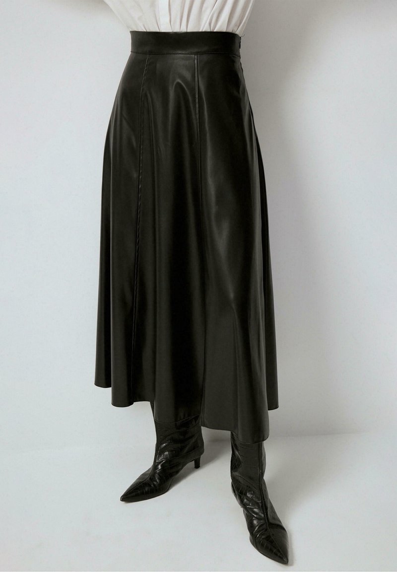 Jupe midi en cuir noire taille haute en forme trapèze, associée à des bottes noires à talons pointus et un top blanc rentré.