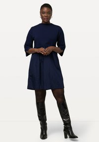 Navyblaues Kleid mit dreiviertel Ärmeln, ausgestelltem Design und hohem Halsausschnitt, kombiniert mit knielangen schwarzen Stiefeln. Glatte Textur.