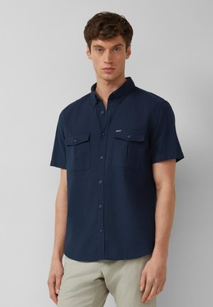 Mann trägt ein marineblaues kurzärmeliges Hemd mit zwei Brusttaschen und steht vor einem einfarbigen hellgrauen Hintergrund.