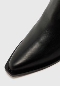 Botín negro de cuero con puntera afilada, textura suave, suela de perfil bajo y diseño angular, que enfatiza una silueta estilizada.