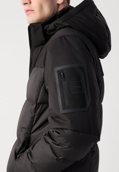 Veste isolée noire avec capuche, dotée d'une poche zippée sur la manche gauche et d'une texture lisse et brillante. Coutures renforcées visibles.
