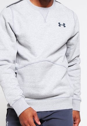 Mand iført en lysegrå Under Armour sweatshirt og mørkeblå bukser, med hænderne afslappet langs siderne mod en ensfarvet baggrund.