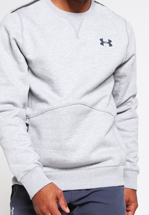 Homme portant un sweat-shirt Under Armour gris clair et un pantalon bleu marine, les mains détendues le long du corps sur un fond uni.