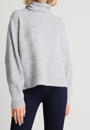 Femme portant un pull à col roulé à côtes gris clair à manches longues et un pantalon sombre ajusté sur un fond uni.