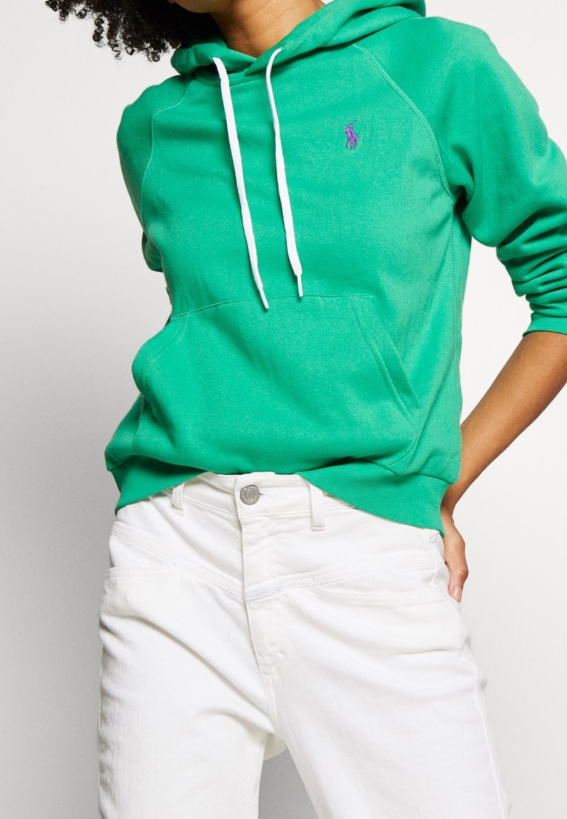 Personne portant un sweat à capuche vert avec des cordons blancs et un pantalon blanc, la main sur la hanche, contre un fond clair et uni.