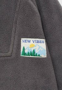 Sivi flis materijal s ušivenim kvadratnim zakrpom na kojoj piše "NOVE VIBRE", planine, žute sunce i zelena stabla. Mekana tekstura i ležeran dizajn.