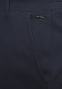 Navyblaue maßgeschneiderte Hose aus strukturiertem Stoff, mit einem kleinen Metalletikett, seitlichen Taschen und einem glatten Bunddesign.