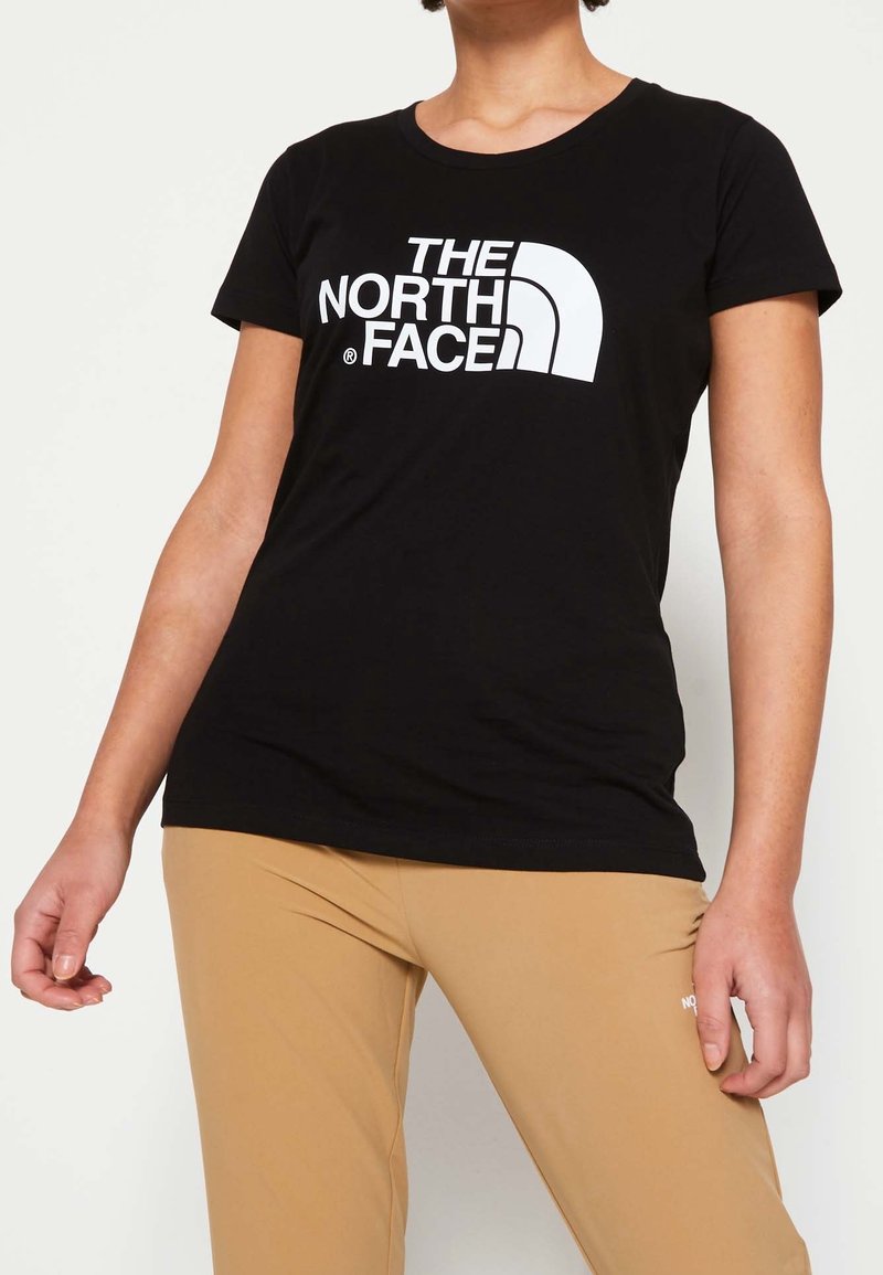 Žena v černém tričku s krátkým rukávem, na kterém je bílé logo "THE NORTH FACE", a v béžových kalhotách na jednobarevném pozadí.