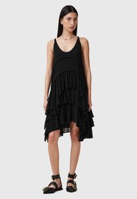 Vestido negro sin mangas con volantes en capas de diferentes longitudes y un escote redondeado. Llevado con sandalias negras de tiras y accesorios minimalistas.