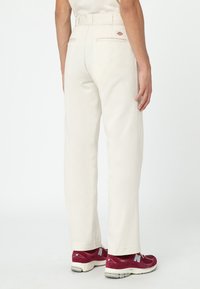 Person in off-white, geradlinigen Hosen mit Gesäßtaschen und maroonfarbenen New Balance Sneakers, steht vor einem schlichten weißen Hintergrund.